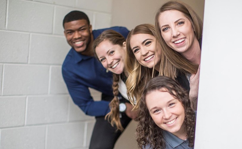 5 co-workers looking around a corner and posing and smiling for a picture